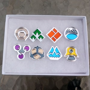 May include: A set of eight metallic enamel pins arranged in two rows inside a white rectangular box. The pins feature various geometric shapes and colors, including green, orange, blue, purple, yellow, and silver accents with distinct patterns.