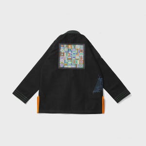 Könnte beinhalten: Schwarze Jacke mit einem farbenfrohen geometrischen Aufnäher auf dem Rücken. Die Jacke hat lange Ärmel, einen orangefarbenen Saum am unteren Rand und ein blau-weißes Design am rechten Ärmel. Die Jacke wird vor weißem Hintergrund gezeigt.