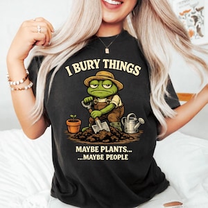 Può includere: T-shirt nera con la scritta "I BURY THINGS" sopra un cartone animato di una rana con cappello e tuta, che tiene una pala. Sotto la rana c'è la scritta "MAYBE PLANTS... ...MAYBE PEOPLE". Sono presenti anche un annaffiatoio e un vaso.