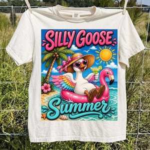 Può includere: T-shirt color crema con una grafica colorata. Raffigura un'oca con occhiali da sole e un cappello di paglia, che galleggia su un galleggiante a forma di fenicottero rosa. Il testo "SILLY GOOSE SUMMER" è visualizzato.