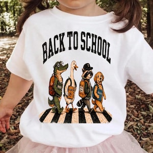 Può includere: Una t-shirt bianca con la grafica di un alligatore, un'oca, un procione e un cane che camminano sulle strisce pedonali. Gli animali indossano zaini e portano libri con le scritte Math, Science e English. Il testo BACK TO SCHOOL è stampato sopra.