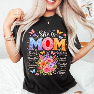 Può includere: T-shirt nero con la scritta colorata "She is MOM" e un design floreale. La maglietta include le parole "Strong", "Never Alone", "Victorious", "Greatly Loved", "Capable", "Beautiful" e "Chosen".
