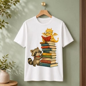 Può includere: Una t-shirt bianca a maniche corte con una stampa grafica di un drago giallo con occhiali che legge un libro rosso, appollaiato su un'alta pila di libri colorati, con un piccolo procione marrone in piedi accanto alla pila di libri.