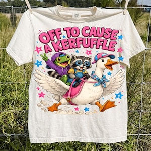 Può includere: T-shirt bianco sporco con una grafica stravagante. Il design presenta una rana, un procione e un'oca, con il testo "OFF TO CAUSE A KERFUFFLE" in rosa e blu. I personaggi cavalcano l'oca, circondati da stelle.