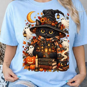 Può includere: Una t-shirt azzurro chiaro con la grafica di un gatto nero con un cappello da strega, circondato da fantasmi, zucche, foglie autunnali, una luna crescente e una pila di libri intitolati Ghost Stories, Magic & Potions e Book of Spells.