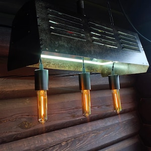 Può includere: Un lampadario industriale in metallo nero con tre lampadine in stile Edison. L'apparecchio rettangolare ha fessure orizzontali e una striscia luminosa. Le lampadine sono sospese all'apparecchio con supporti cilindrici neri.