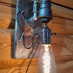 May include: Industrial-style wall sconce featuring a vintage Edison bulb. The light fixture is constructed from metal pipes and fittings, mounted on a weathered wooden plank. The bulb emits a warm, inviting glow, perfect for rustic decor.