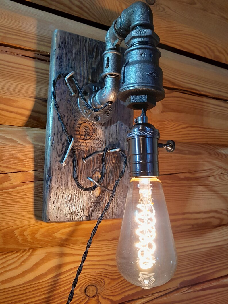 May include: Industrial-style wall sconce featuring a vintage Edison bulb. The light fixture is constructed from metal pipes and fittings, mounted on a weathered wooden plank. The bulb emits a warm, inviting glow, perfect for rustic decor.