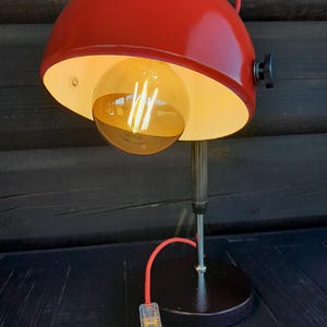Può includere: Una lampada da scrivania rossa con un paralume arrotondato e una grande lampadina a vista. La lampada ha una base nera e un cavo di alimentazione rosso. La lampadina è di colore giallo caldo e il paralume ha una maniglia.