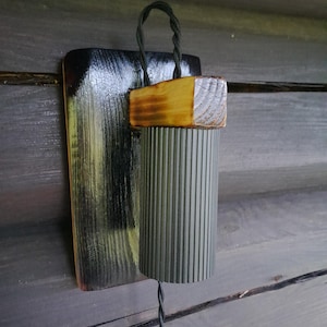 Op de afbeelding: Een handgemaakte houten wandlamp met een donkere afwerking. De lamp heeft een rechthoekige achterplaat, een cilindrische kap met verticale groeven en een houten blok. Een zwart snoer is bevestigd om op te hangen.