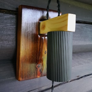 May include: A wall-mounted lamp with a dark green, ribbed cylindrical shade. It is attached to a rectangular wooden base with a natural wood finish. The design includes a twisted black cord and a light-coloured wooden beam.