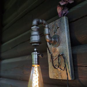 May include: Industrial-style wall sconce featuring a vintage Edison bulb. The fixture is constructed from black metal pipes and fittings, mounted on a weathered wooden board. A decorative lighting element for home decor.