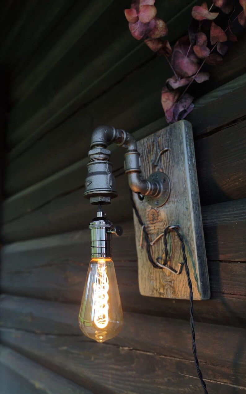 May include: Industrial-style wall sconce featuring a vintage Edison bulb. The fixture is constructed from black metal pipes and fittings, mounted on a weathered wooden board. A decorative lighting element for home decor.