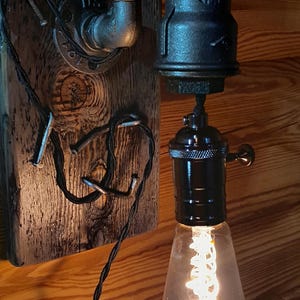 May include: Industrial-style wall sconce featuring a black metal pipe design. The fixture includes a vintage-style Edison bulb with a visible filament, mounted on a wooden plank. The wiring is visible, adding to the rustic aesthetic.