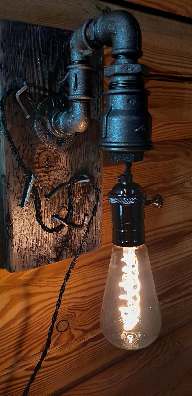 May include: Industrial-style wall sconce featuring a black metal pipe design. The fixture includes a vintage-style Edison bulb with a visible filament, mounted on a wooden plank. The wiring is visible, adding to the rustic aesthetic.