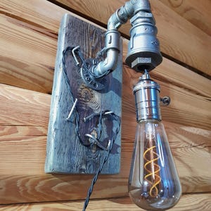 May include: Industrial-style wall sconce featuring a gray metal pipe design mounted on a weathered wooden plank. The light fixture includes a decorative Edison bulb with a warm, orange filament. The electrical cord is black.