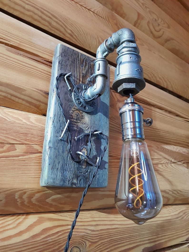 May include: Industrial-style wall sconce featuring a gray metal pipe design mounted on a weathered wooden plank. The light fixture includes a decorative Edison bulb with a warm, orange filament. The electrical cord is black.