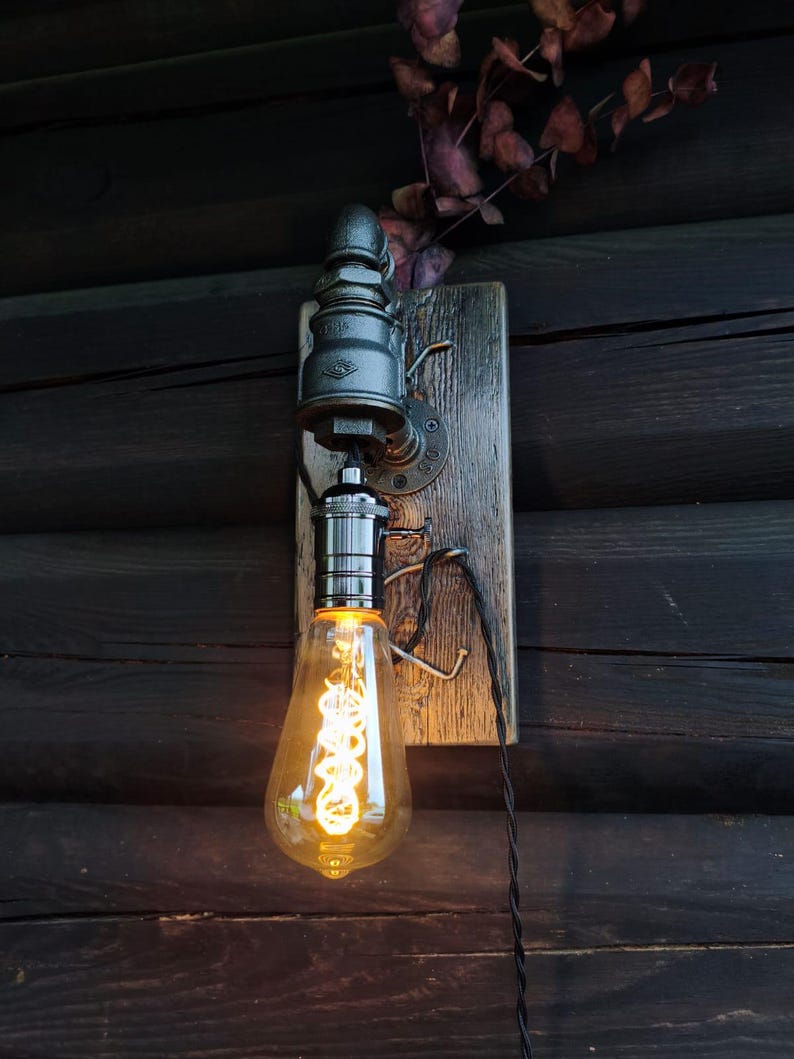 May include: Industrial-style wall sconce featuring a vintage Edison bulb with a warm glow. The light fixture is mounted on a weathered wooden board with metal piping and a black electrical cord. The design incorporates rustic and steampunk elements.