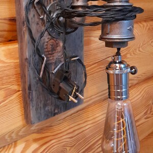 May include: Industrial-style wall sconce featuring a vintage Edison bulb. The fixture is constructed from metal pipes and fittings, mounted on a weathered wooden plank. The bulb has a visible filament and is encased in clear glass.