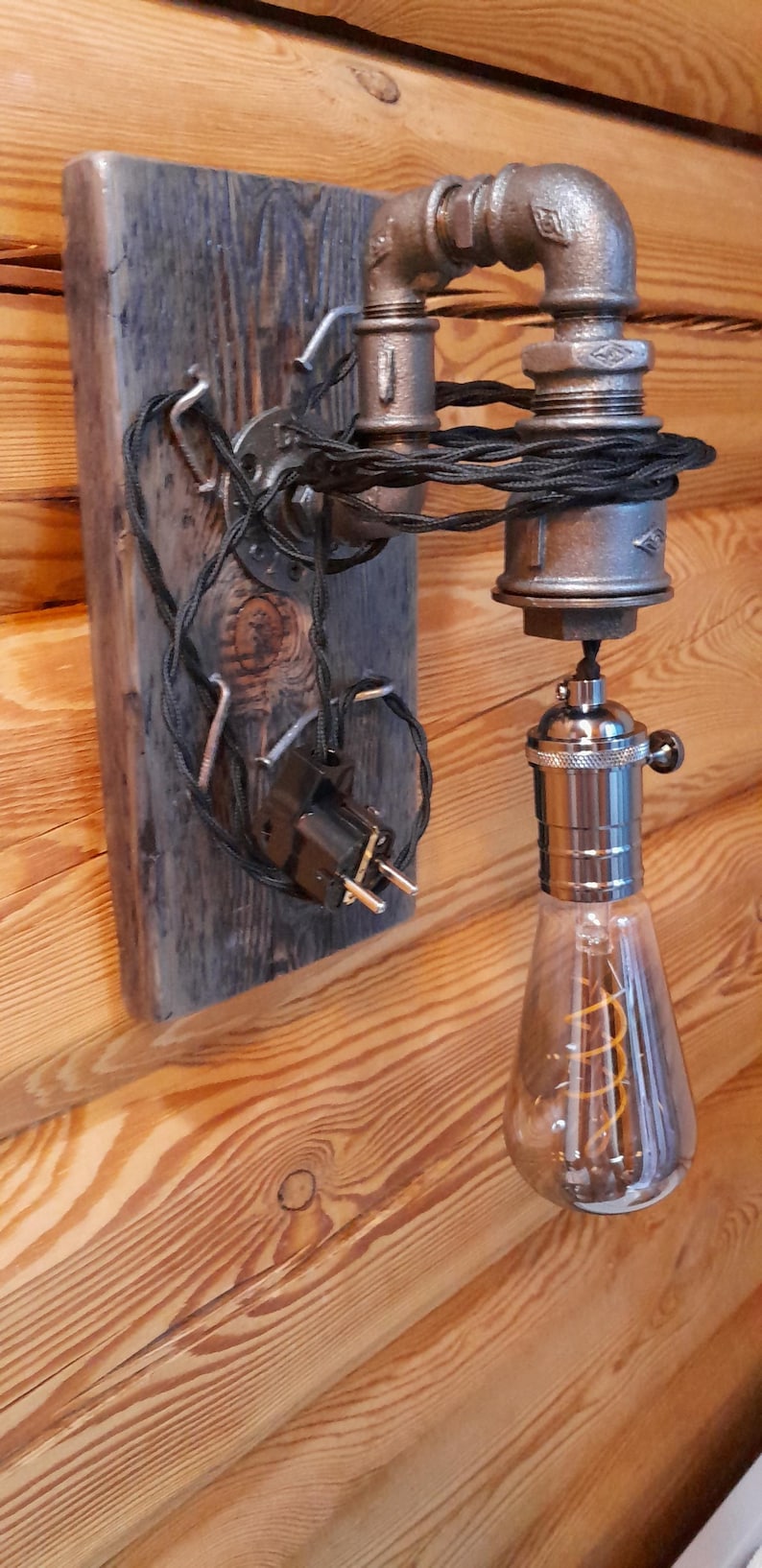 May include: Industrial-style wall sconce featuring a vintage Edison bulb. The fixture is constructed from metal pipes and fittings, mounted on a weathered wooden plank. The bulb has a visible filament and is encased in clear glass.