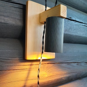 May include: A contemporary wall light featuring a light wood mounting plate and a black, ribbed metal shade. The light has a twisted black cable and casts a warm, ambient glow. The design is simple and suitable for various interior styles.