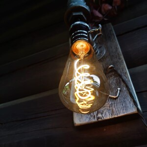 May include: A vintage-style light bulb with a warm, glowing filament. The bulb is teardrop-shaped and encased in clear glass. It is mounted on a weathered wooden plank with metal hardware. The overall aesthetic is rustic and industrial.