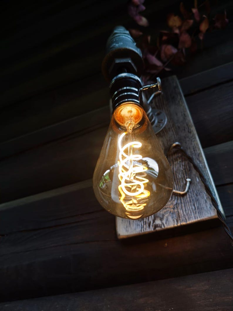 May include: A vintage-style light bulb with a warm, glowing filament. The bulb is teardrop-shaped and encased in clear glass. It is mounted on a weathered wooden plank with metal hardware. The overall aesthetic is rustic and industrial.