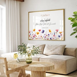 Puede incluir: Una impresión artística enmarcada con borde dorado, con caligrafía árabe y coloridas flores de acuarela. La impresión cuelga sobre un sofá beige en un salón moderno y luminoso. A la derecha hay una gran planta en maceta.