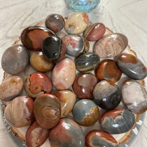 May include: A collection of polished stones in various colors, including red, brown, gray, and cream. The stones are smooth and oval-shaped, displayed on a wooden slice. A decorative glass vase is in the background.