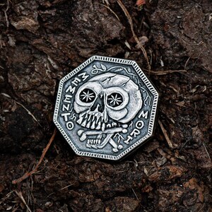 May include: A silver-toned, octagonal-shaped coin with a skull design and the words "Memento Mori" inscribed around the edge. The coin is lying on a dark brown, textured surface.