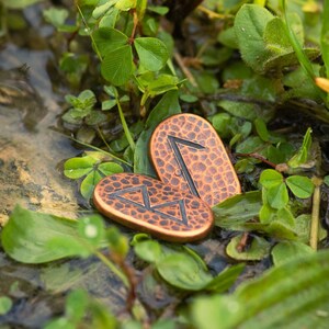 May include: Two copper-colored metal runes with black markings, lying on a bed of green clover leaves. The runes are shaped like rounded rectangles with a textured surface.