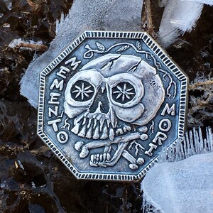 May include: A silver octagonal coin with a skull and crossbones design. The skull has a cracked face and the text "Memento Mori" is inscribed around the edge.
