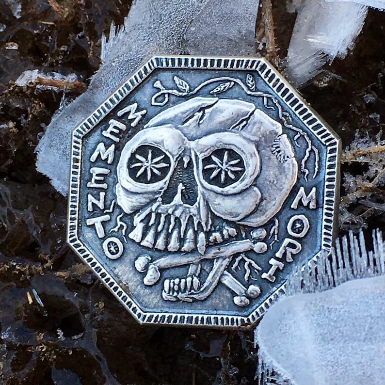 May include: A silver octagonal coin with a skull and crossbones design. The skull has a cracked face and the text "Memento Mori" is inscribed around the edge.