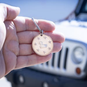 May include: A silver keychain with a round, silver charm featuring a duck silhouette and a star design. The charm is held in a hand against a blurred background of a white Jeep.