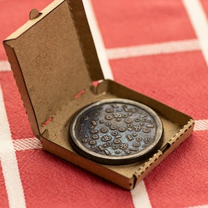 Pode incluir: Um token de pizza redondo de metal com um design de vários ingredientes de pizza em uma caixa de pizza de papelão.