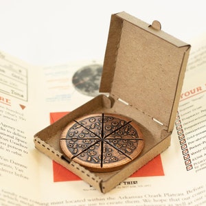 May include: A copper-colored pizza-shaped coin, divided into eight slices, rests inside a small cardboard pizza box. The coin features detailed toppings. The box is open, revealing the coin on a printed background.