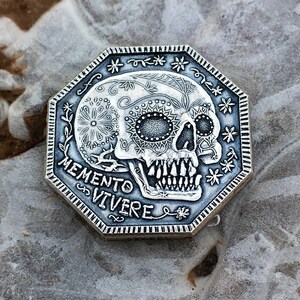 May include: A silver octagonal coin with a detailed sugar skull design and the words "Memento Vivere" inscribed around the skull.