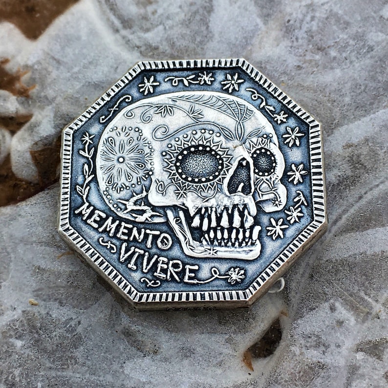 May include: A silver octagonal coin with a detailed sugar skull design and the words "Memento Vivere" inscribed around the skull.
