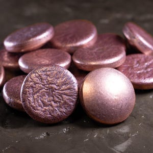 Può includere: Una collezione di perline piatte e rotonde color rame. Alcune perline hanno una superficie strutturata, mentre altre sono lisce. Le perline sono sparse su una superficie scura e strutturata, creando un'estetica metallica e terrosa. Ideale per la creazione di gioielli.