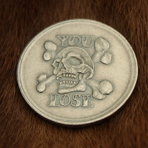 May include: A silver-colored coin with a skull and crossbones design. The coin reads "YOU LOSE" in a stylized font.