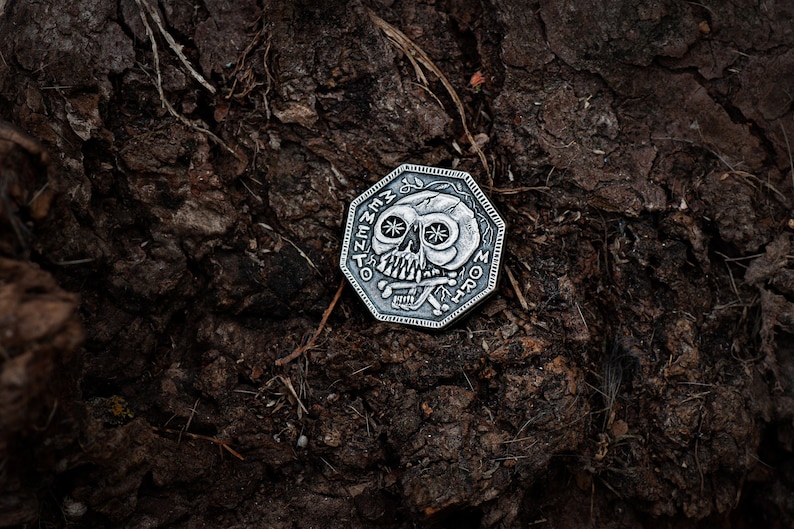 May include: A silver-toned, octagonal-shaped coin with a skull design and the words "Memento Mori" inscribed around the edge. The coin is lying on a dark brown, textured surface.