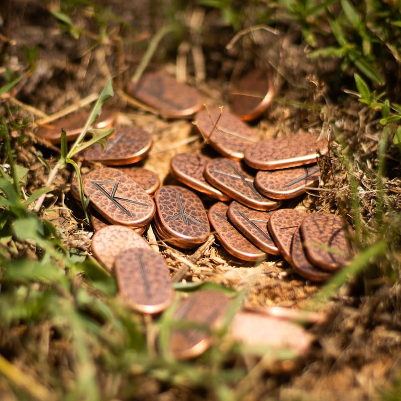 May include: A collection of copper-colored metal runes with etched symbols, scattered on a bed of green grass and brown dirt.