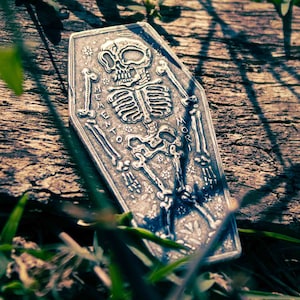 Puede incluir: Un colgante de plata en forma de ataúd con un esqueleto en su interior. El colgante tiene las palabras "Memento Mori" grabadas.