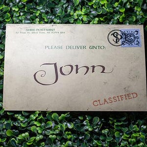 May include: A rectangular tan envelope with the text SHIRE-POST-MINT, 82 Main St, West Fork, AR 72774 USA, PLEASE DELIVER UNTO: John, and CLASSIFIED printed on the front. A blue and black circular stamp is located in the top right corner of the envelope.
