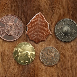 May include: Five different metal coins with various designs. The coins are on a brown fur surface. One coin features a horse, another a bird, another a leaf with text, another a lion, and the last one has an eye with text.