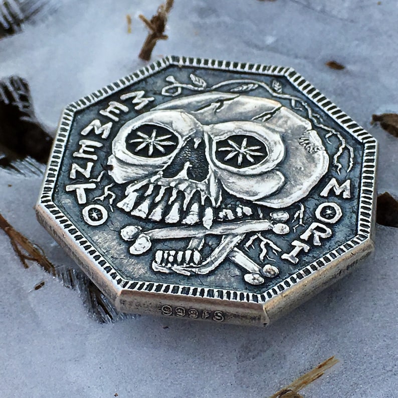 May include: A silver octagonal coin with a skull design. The skull has a cracked face with two star-shaped eyes and a cross bone design. The coin has the words "Memento Mori" inscribed around the edge and "999FS" on the bottom.