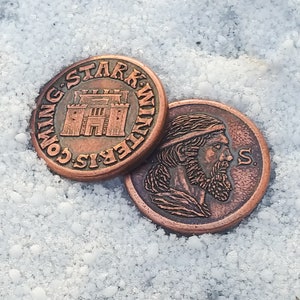 Może przedstawiać: Dwie miedziane monety z motywem Gry o Tron. Jedna moneta przedstawia zamek z tekstem "Winter is Coming. Stark." Druga moneta przedstawia profil mężczyzny z brodą i tekstem "S."