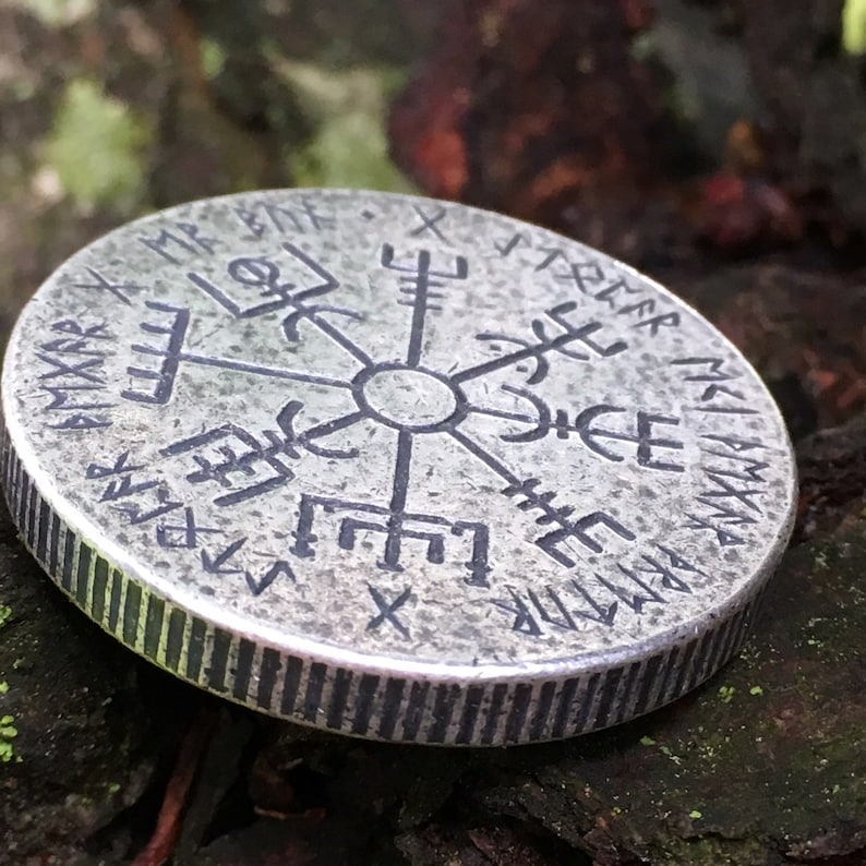 May include: A silver-toned metal coin with a circular design featuring a compass rose with runes around the edge. The runes are in the Old Norse language.