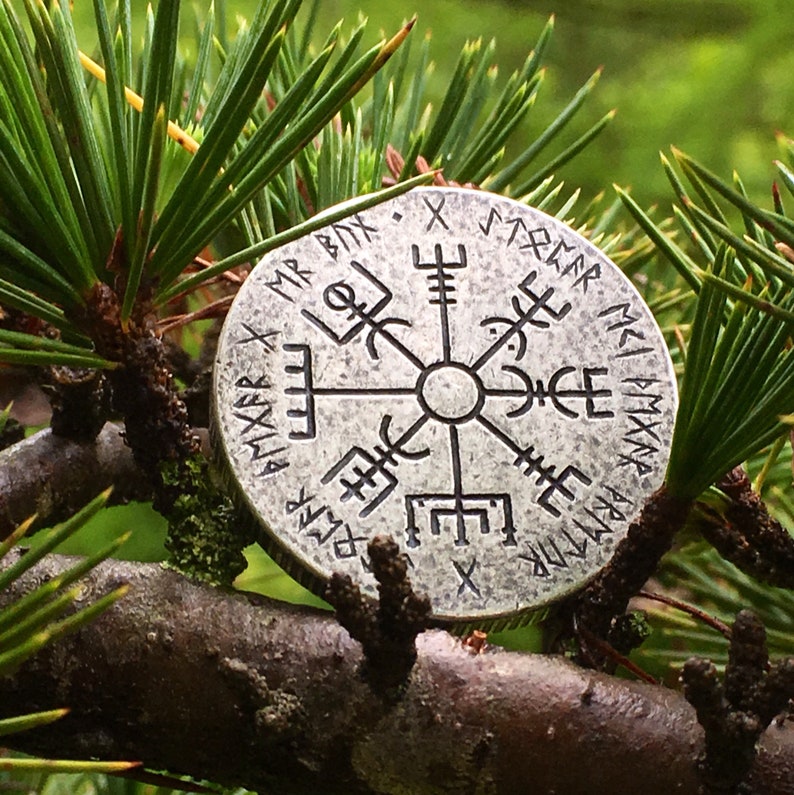 May include: A silver compass with a Vegvisir symbol and runes around the edge. The compass is resting on a branch of a pine tree.