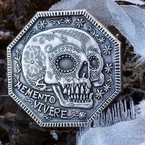 May include: A silver-toned, octagonal coin with a detailed sugar skull design and the words "Memento Vivere" inscribed around the edge.